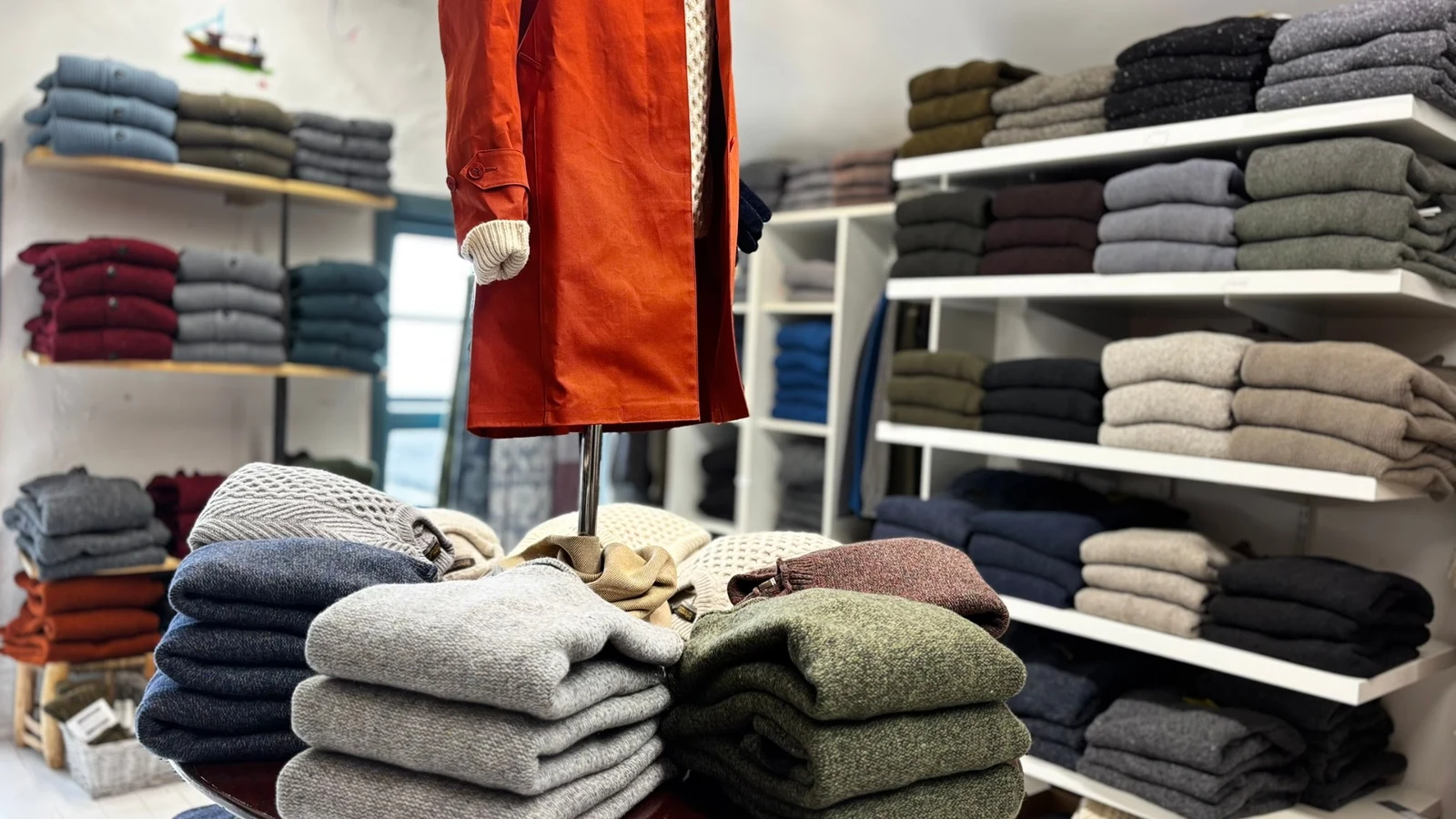 Colourful folded wool knitwear on shelves and a display table beneath an orange coat at Cleo's