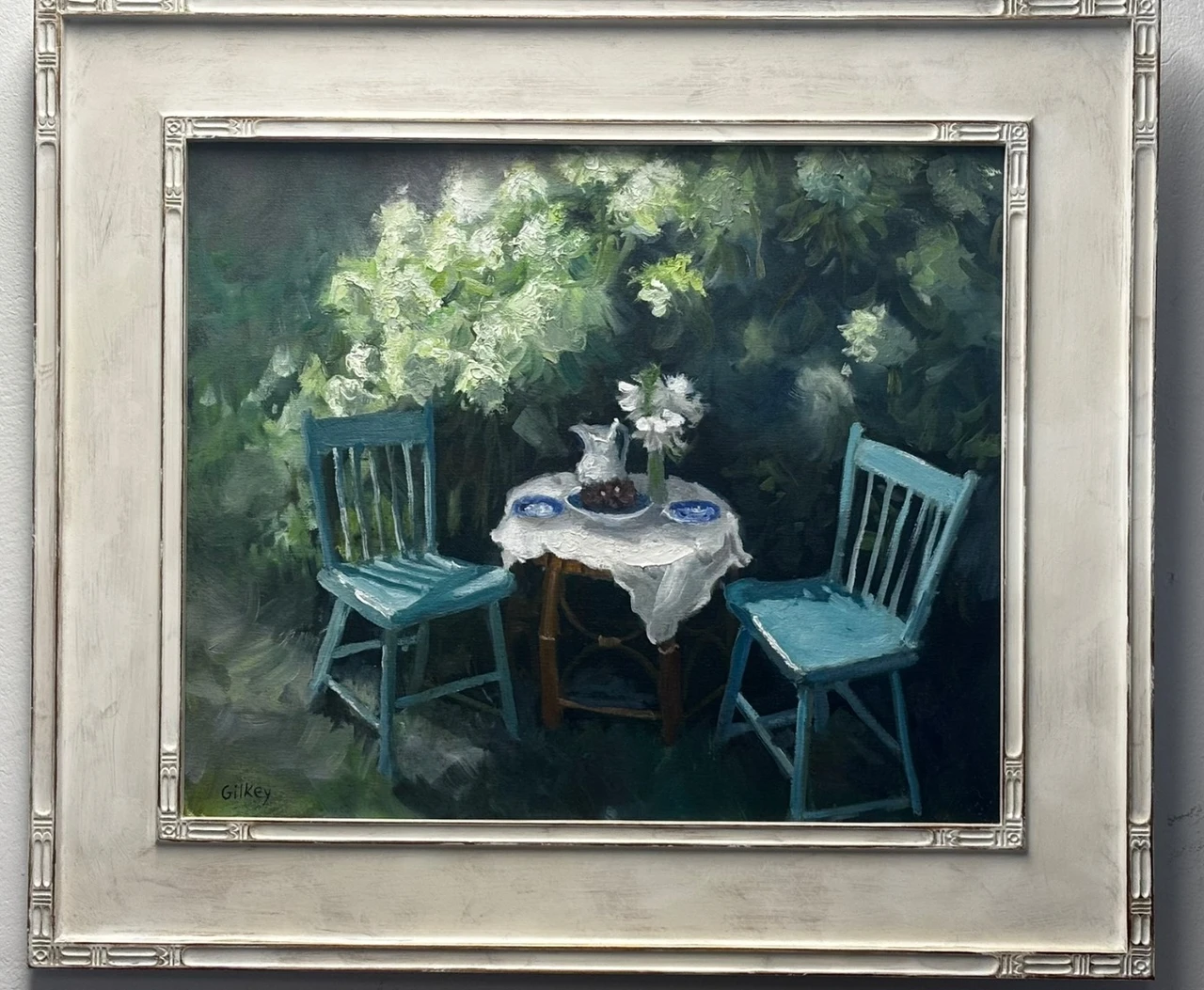 Framed painting of two turquoise chairs beside a small table surrounded by green foliage