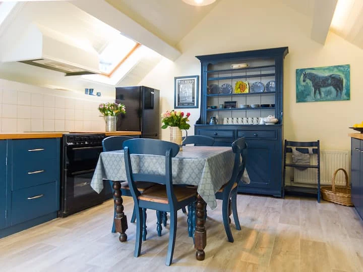 Blue kitchen cabinets and a dresser beside a dining table and chairs in a light-filled accommodation interior